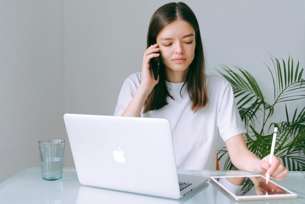 A young woman multitasking with a phone, laptop, and tablet in a bright home office setting.